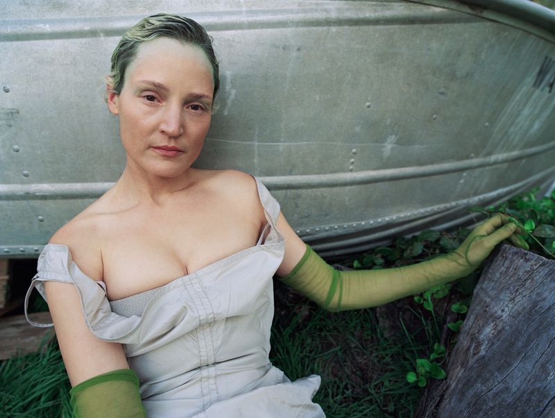 Woman in low-cut dress leans against a boat, wearing long green gloves, surrounded by grass, creating an editorial portrait.
