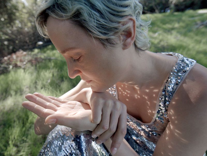 Adult female in a sequined tank dress sitting on grass, looking at her hand with a blurred green and brown background.