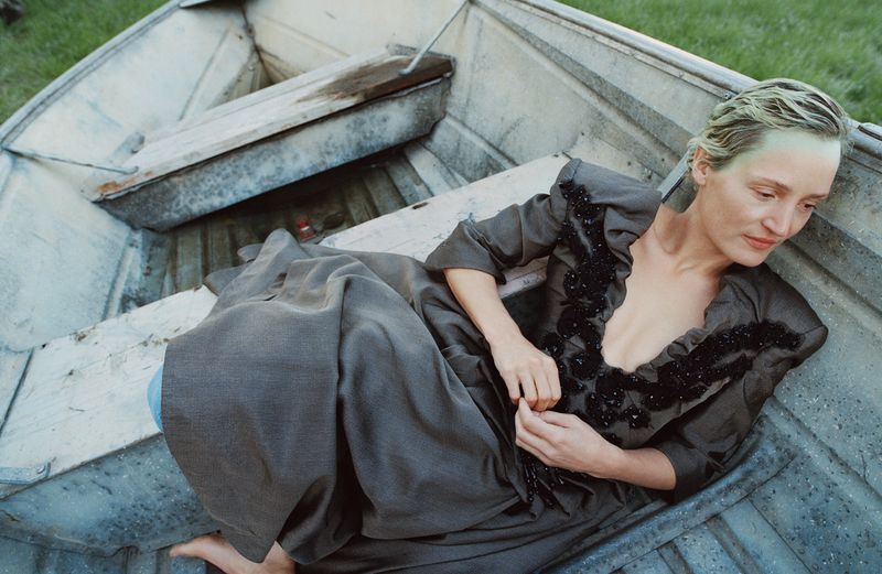 A woman in a grey dress lying in a weathered metal boat on grass with faded green hair, creating a moody outdoor fashion image.