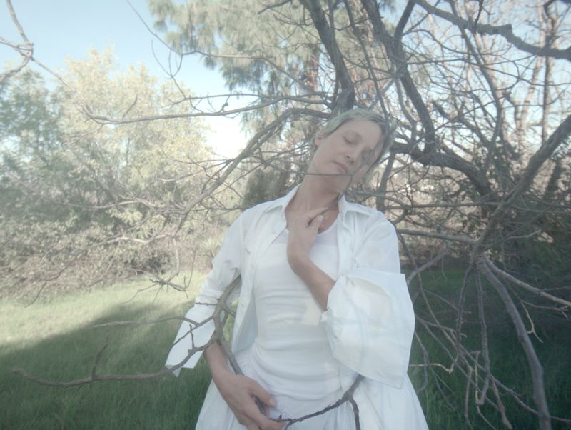 Adult female model with pale skin poses with eyes closed near leafless tree, dressed in white, outdoors on sunny day.