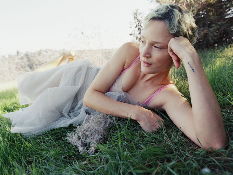 Woman with short hair in a pink top and white dress lies in green grass with a background of trees on a sunny day.