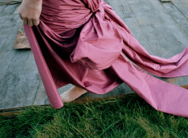 Close-up of actor Vicky Krieps wearing a voluminous pink silk dress over bare feet against a wooden surface and grass.