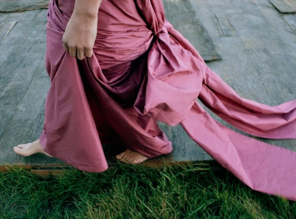 Woman walks barefoot across a wooden surface, wearing a flowing pink gown with a dramatic knot detail, set against a backdrop of green grass.