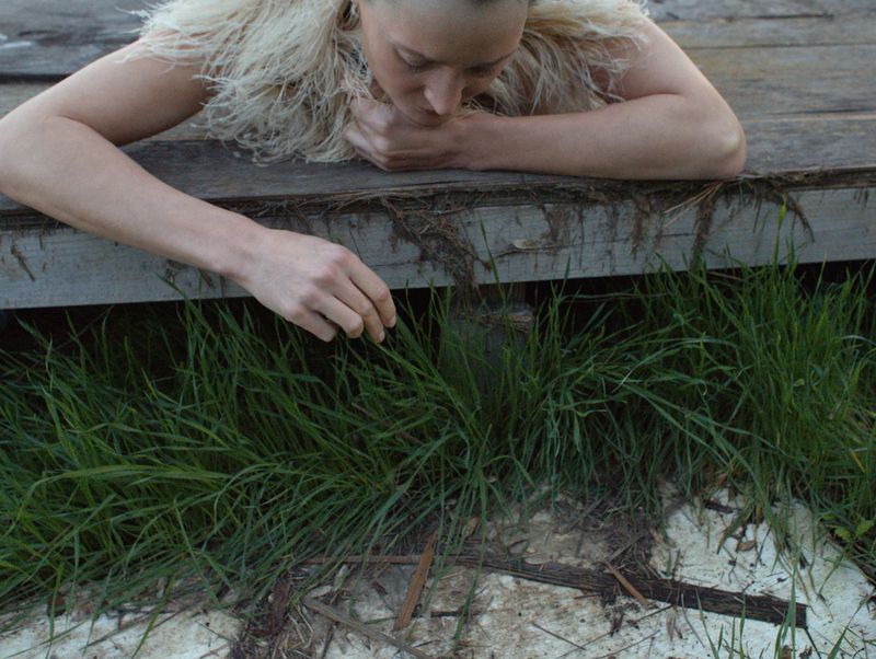 Woman leaning on a wooden structure, looking at green grass; neutral lighting and a shallow depth of field.
