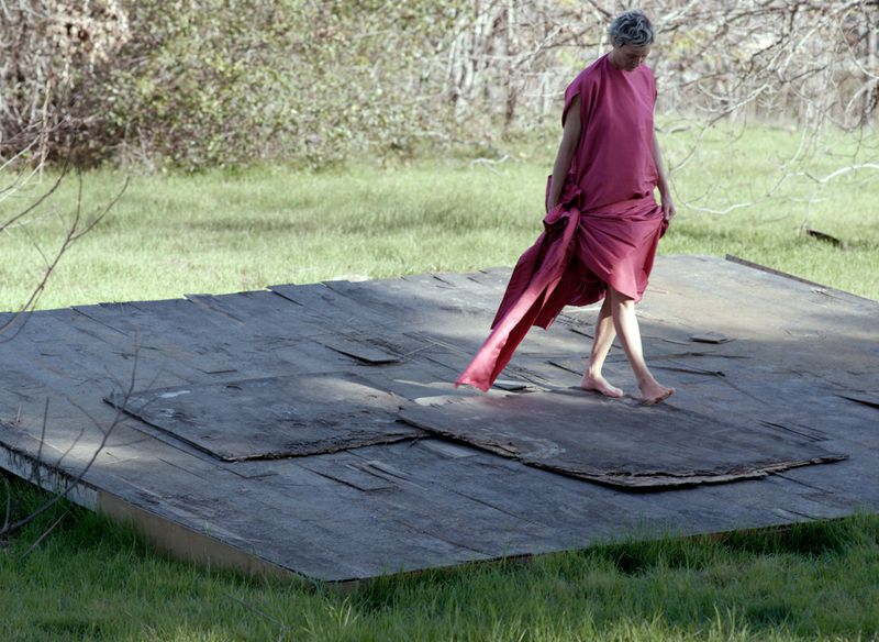 Vicky Krieps in a long pink dress walking barefoot on a weathered wooden platform in a field for AnOther Magazine.