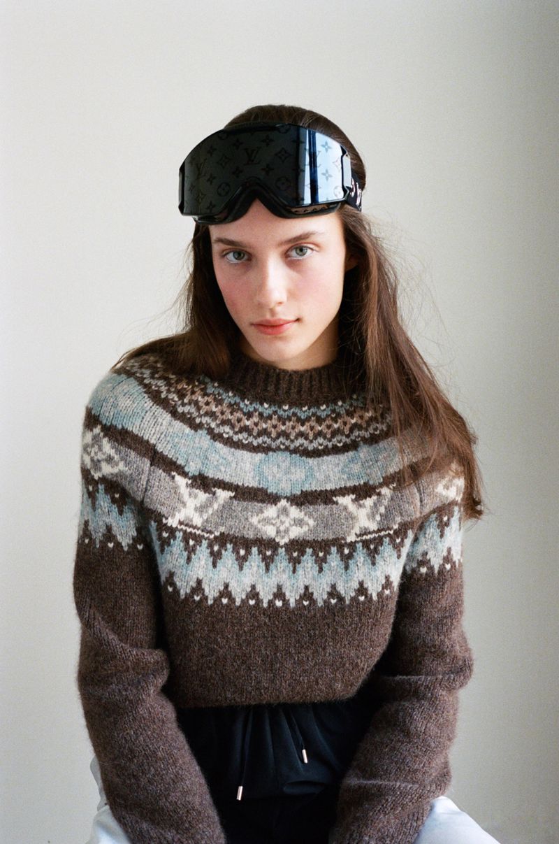 Young woman poses in a patterned sweater wearing ski goggles on her head, indoors against a neutral background in a medium shot.