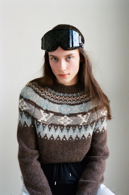 Young woman poses in a patterned sweater wearing ski goggles on her head, indoors against a neutral background in a medium shot.