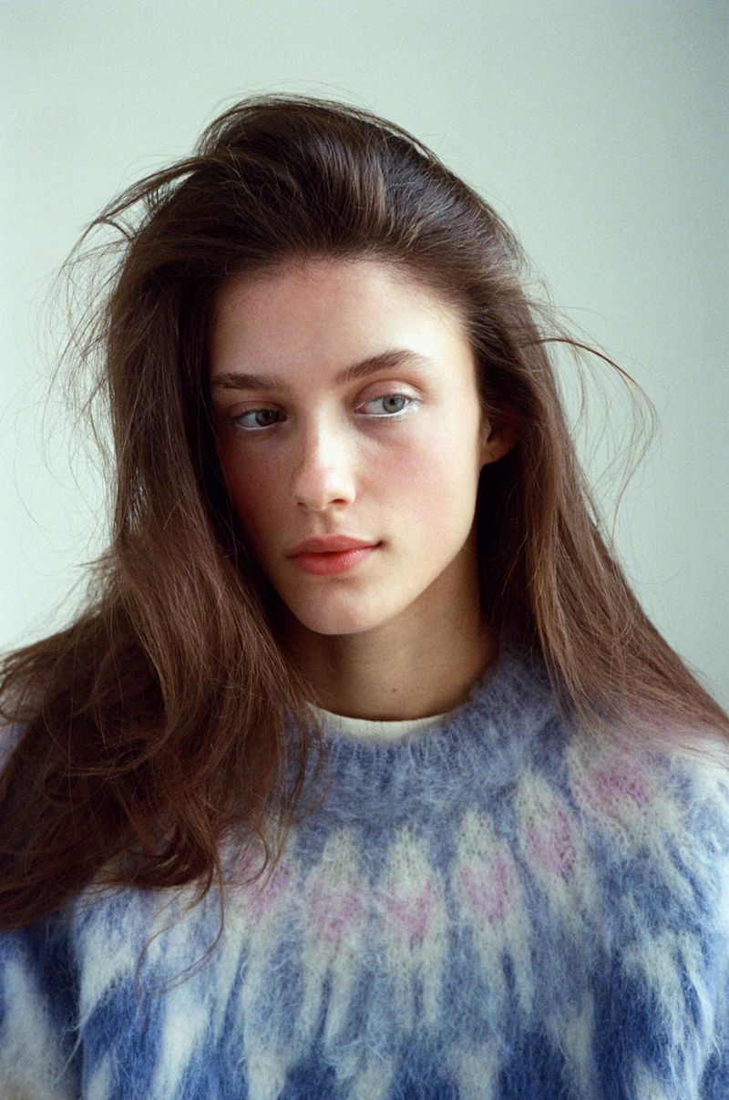 Close-up portrait of a young adult woman with long brown hair wearing a patterned blue and white sweater against a light grey background.