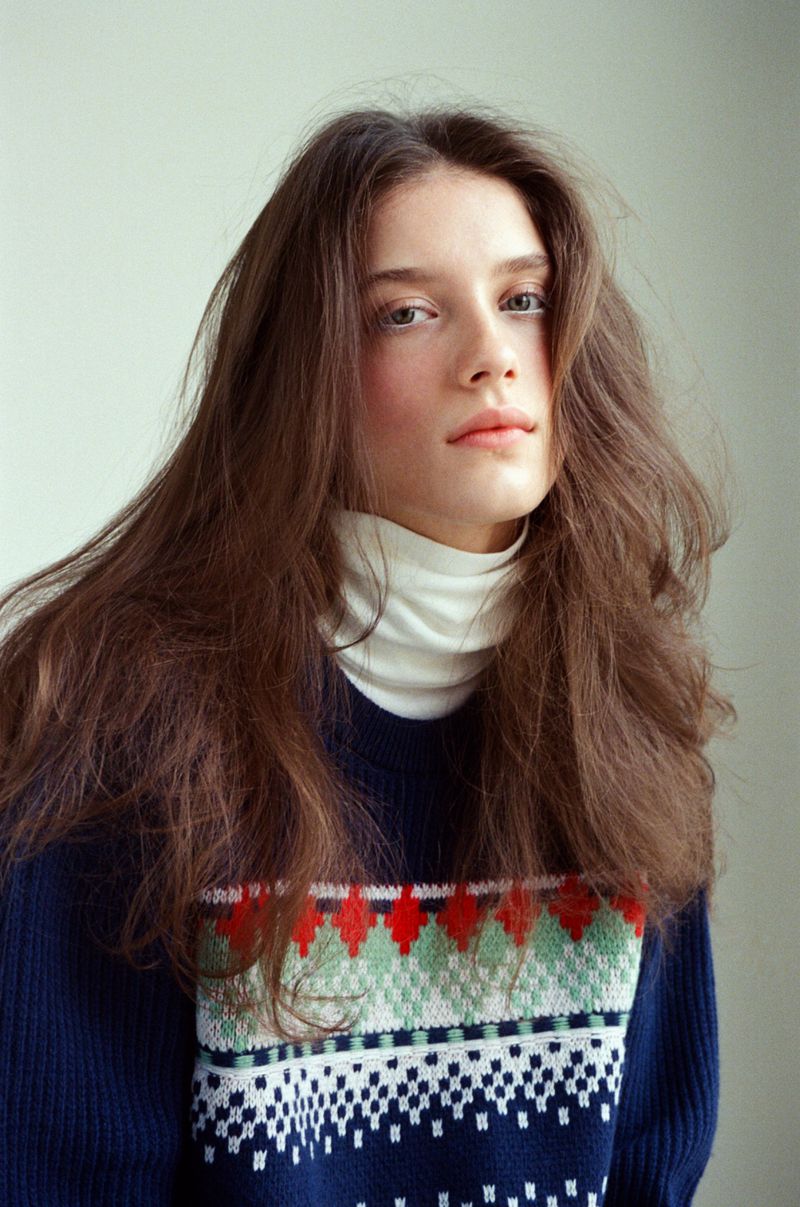 Portrait of a young adult female with long brown hair, wearing a patterned blue sweater and a white turtleneck against a neutral backdrop.
