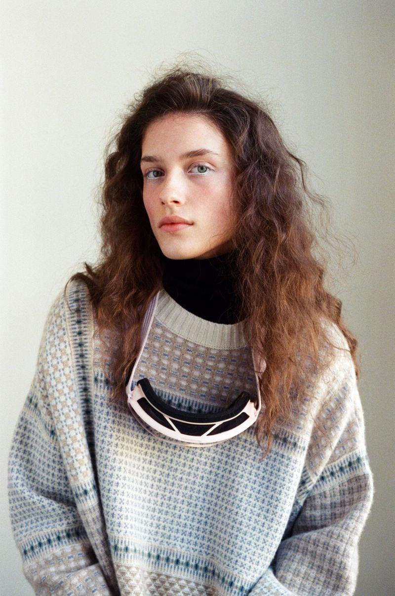 Portrait of a fair-skinned woman with wavy brown hair, wearing a patterned sweater and ski goggles, posing in front of a plain wall.