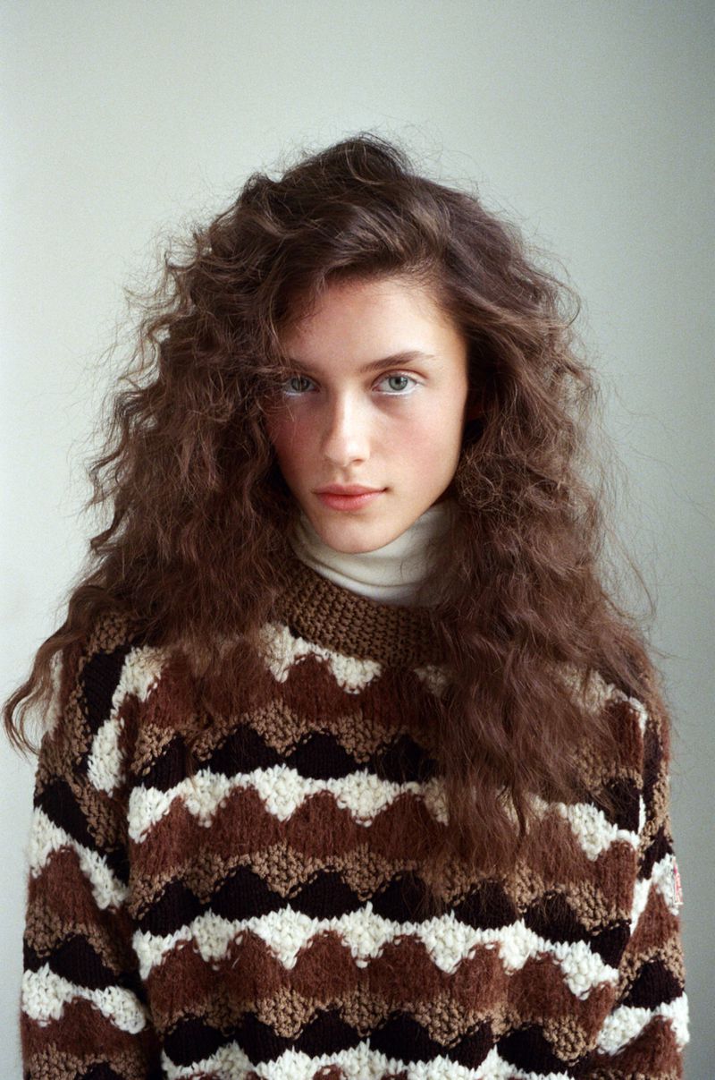 Portrait of a young woman with curly brown hair wearing a patterned sweater against a neutral background, looking directly at the camera.