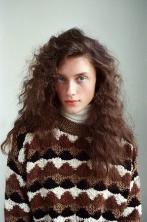 Portrait of a young woman with curly brown hair wearing a patterned sweater against a neutral background, looking directly at the camera.
