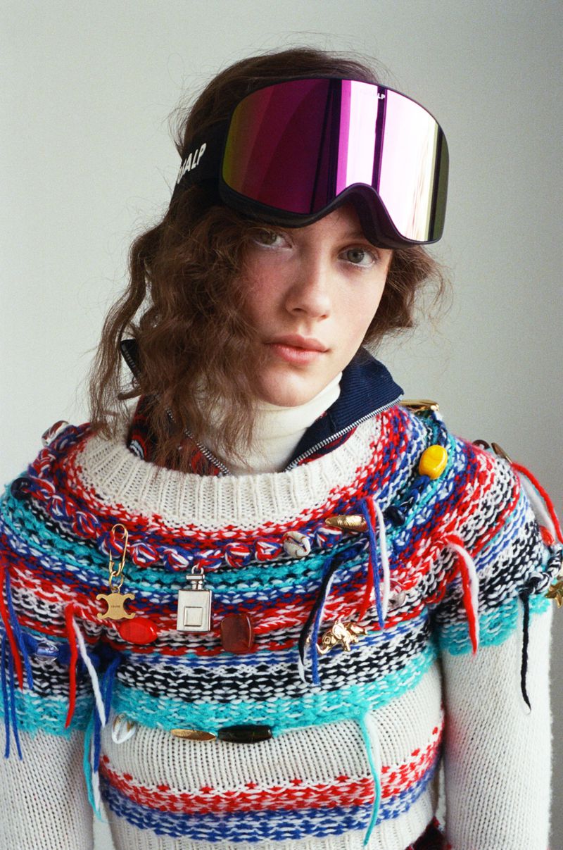 Fashion portrait of a young adult woman wearing colorful knit sweater and ski goggles against a white backdrop in a studio setting.
