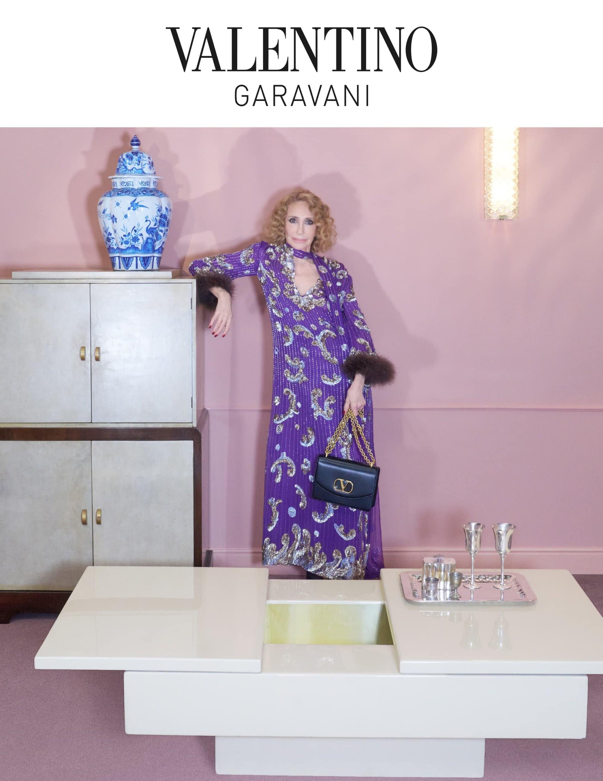 A woman in a purple gown poses next to a cabinet and table in a pink room. She is holding a black purse and wearing brown fur cuffs.