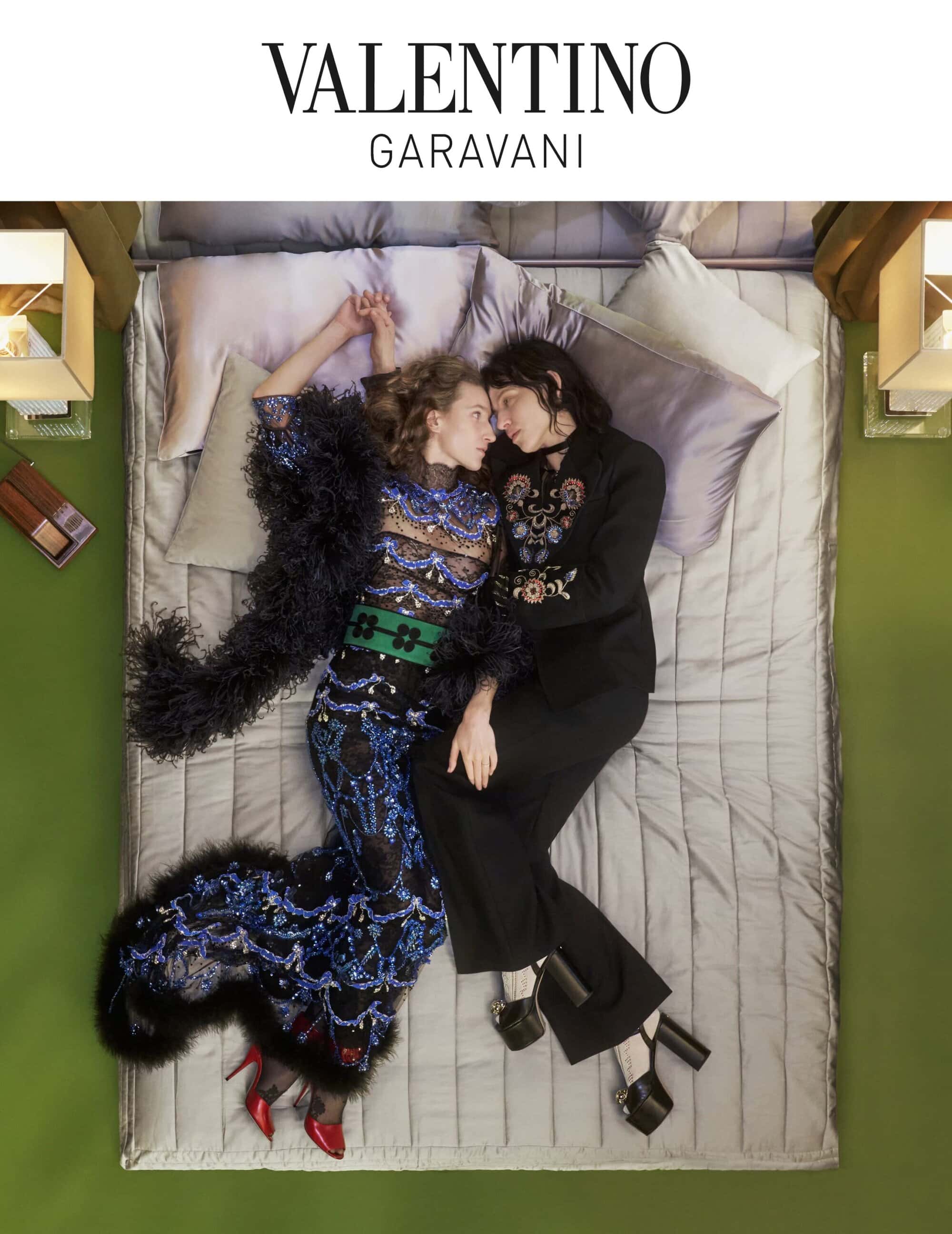 Two women lie on a bed in a fashion editorial, one in a sequined dress with feather boa, the other in a black suit, with a green background.