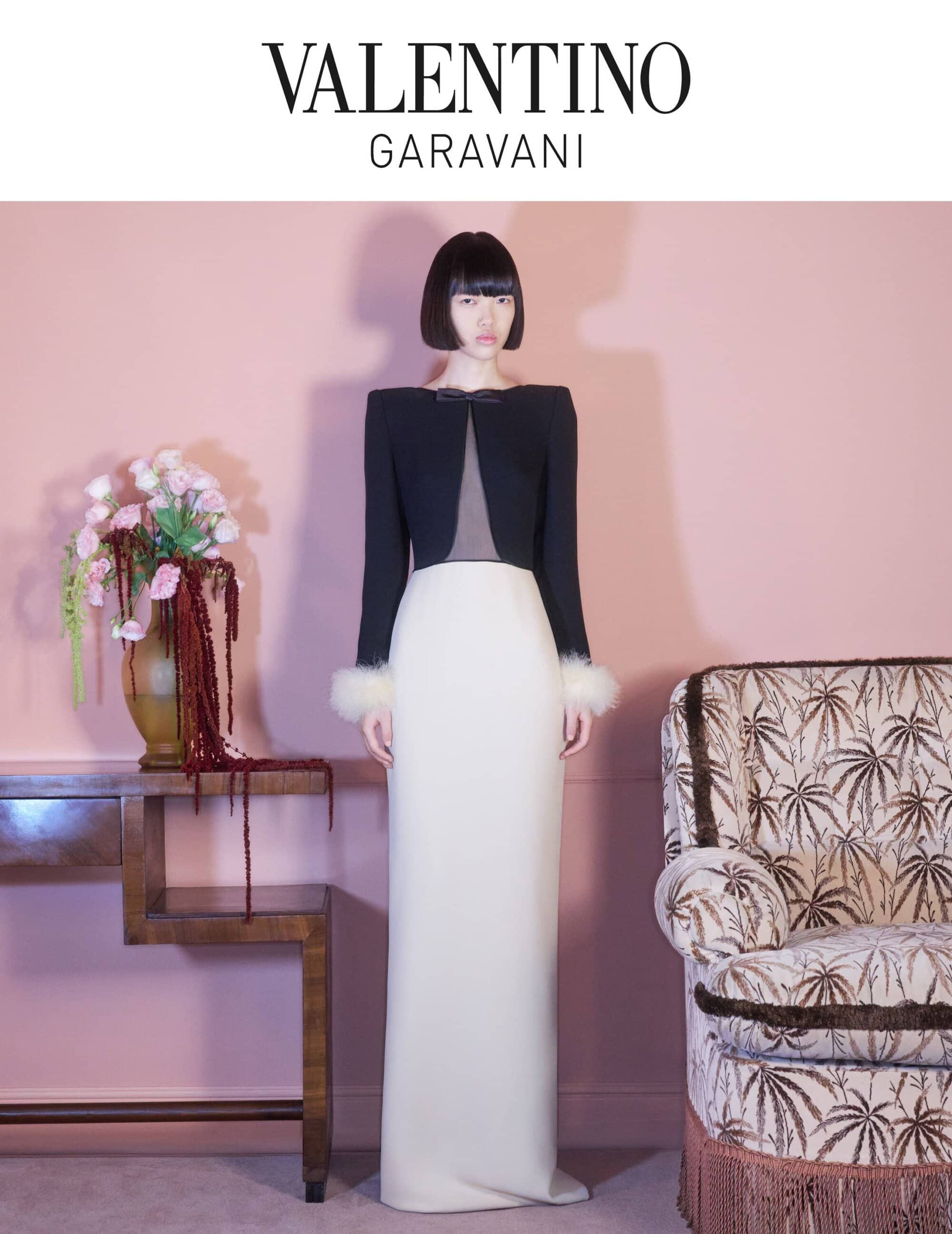 A woman in black jacket and white gown poses in front of a pink wall with floral decorations for a Valentino Garavani advertisement.