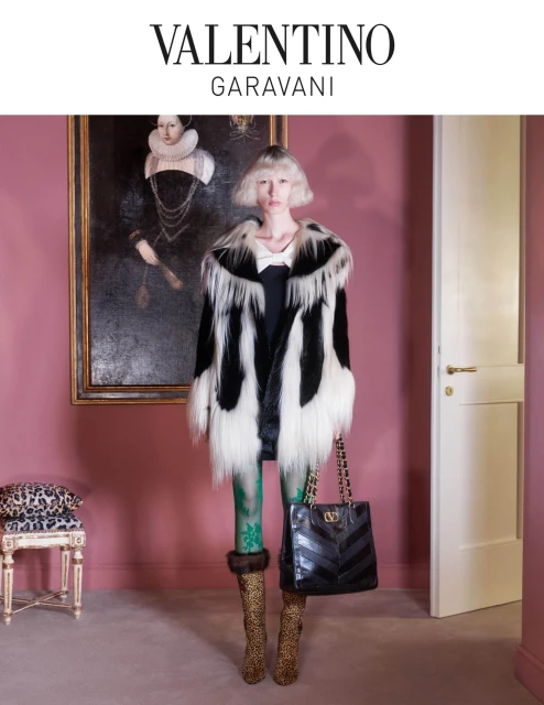 A woman poses in a black and white fur coat, holding a black purse with chain straps, standing in a pink room with a portrait on the wall.