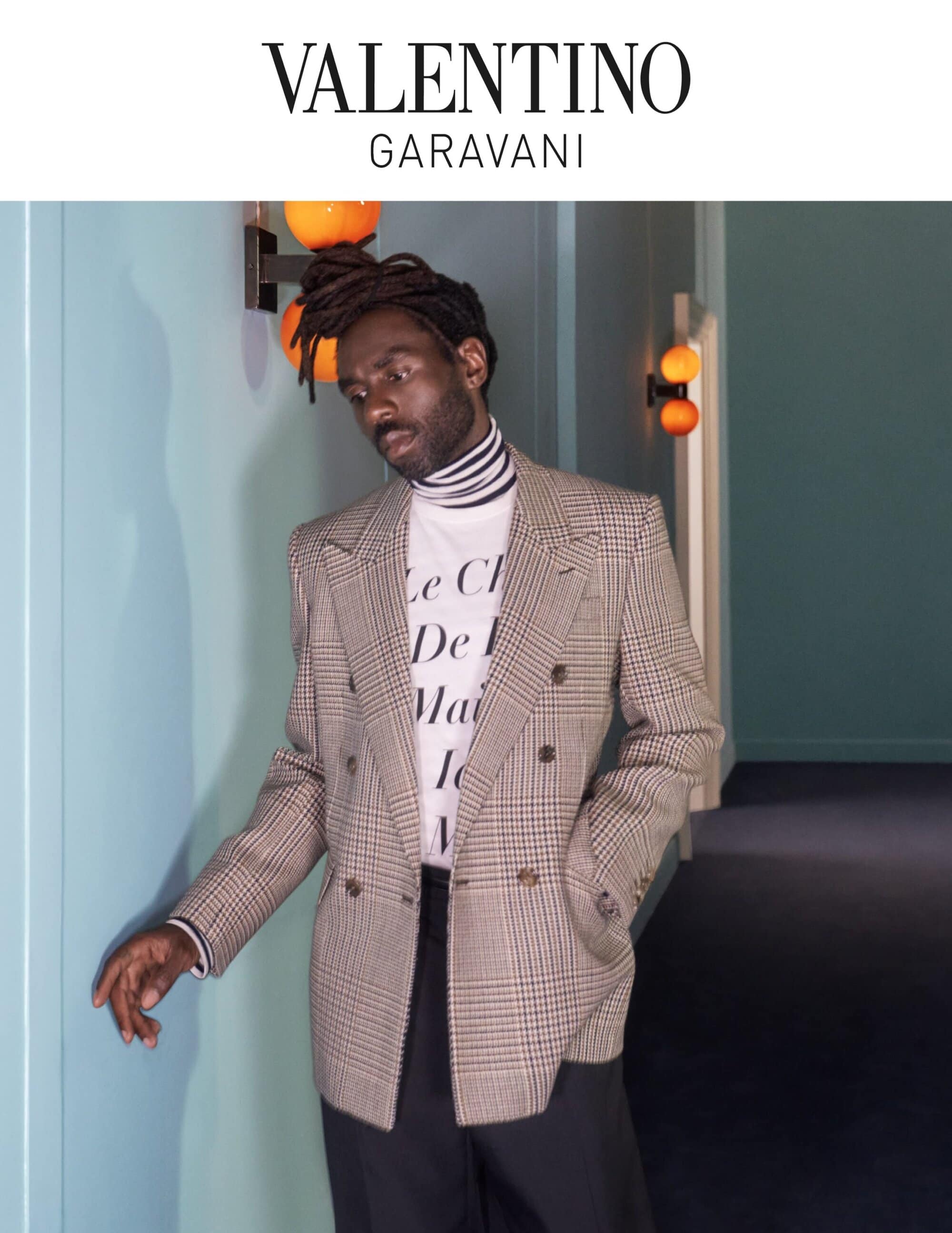 A Black male model poses in a plaid blazer and t-shirt, standing against a light blue wall in an indoor hallway setting with warm lighting.
