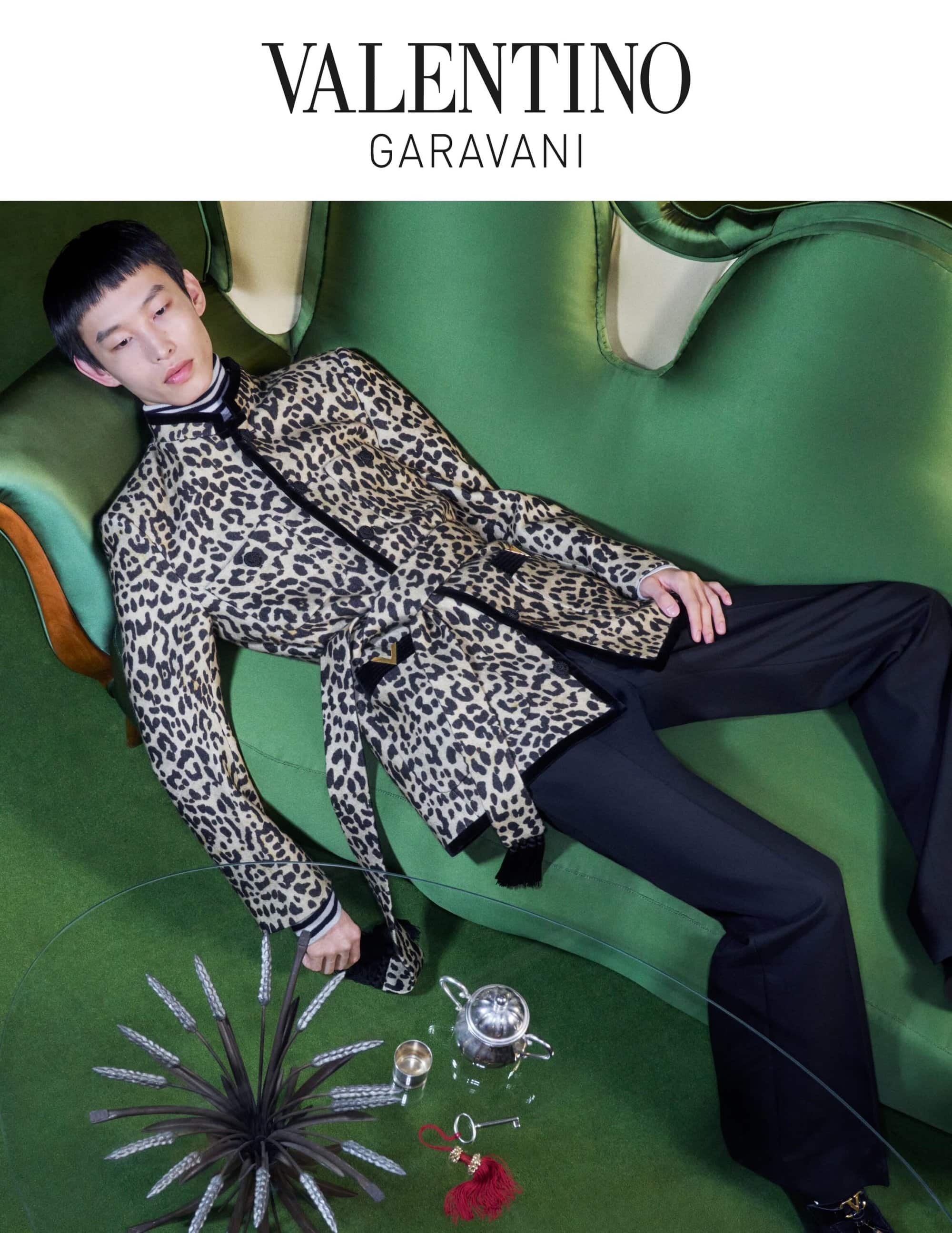Fashion shot featuring an adult male model on a green velvet couch wearing a leopard print coat and black pants, with a modern decor.