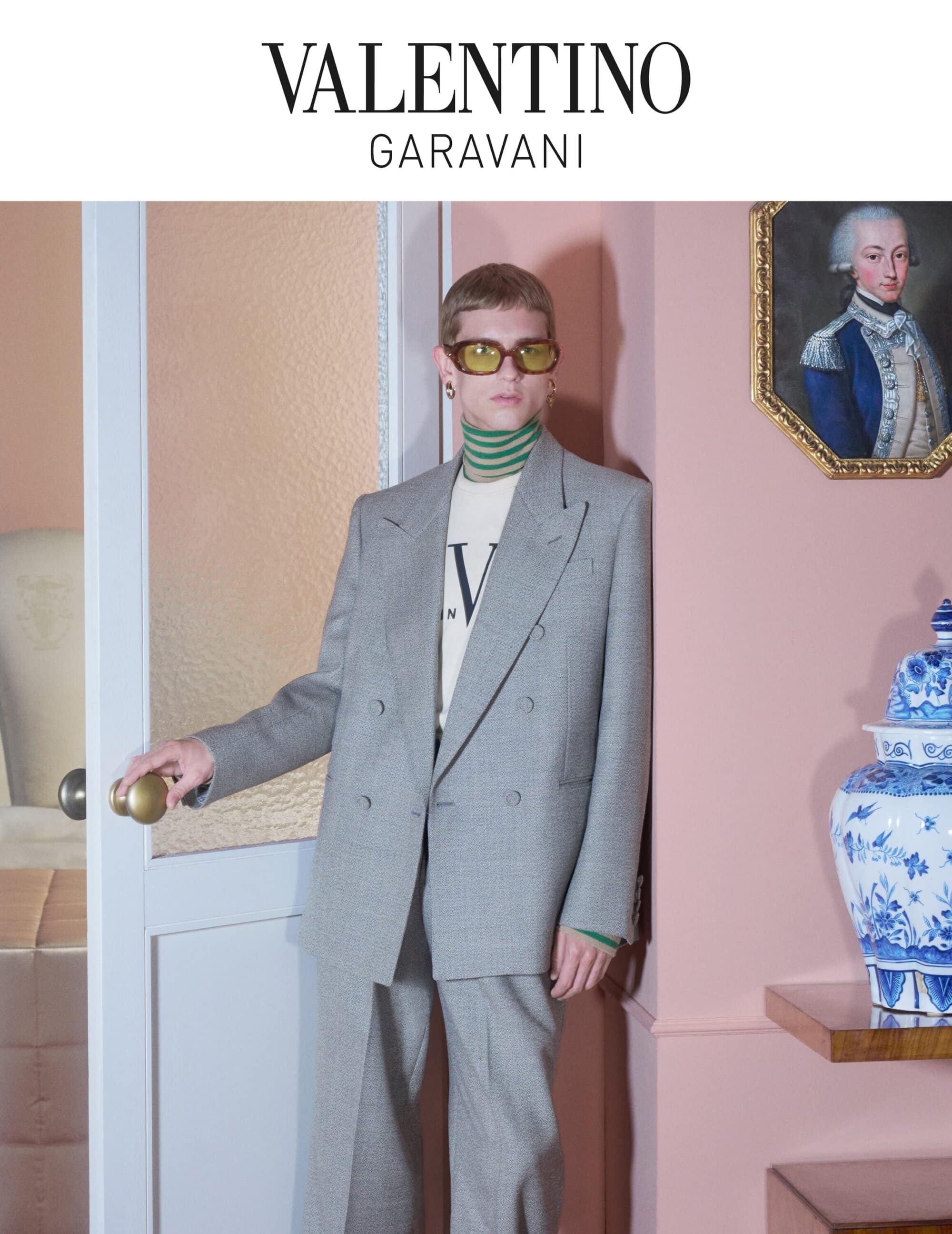 A stylish adult male poses next to a door in a gray suit, wearing yellow sunglasses, turtleneck, and Valentino branded t-shirt indoors.