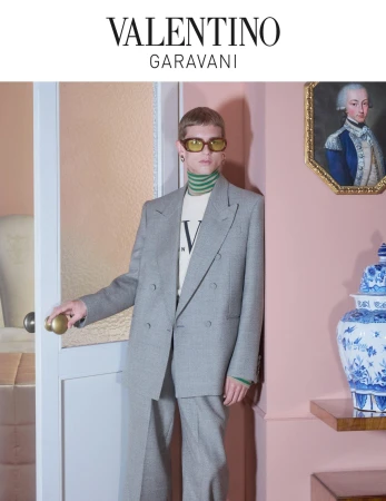 A stylish adult male poses next to a door in a gray suit, wearing yellow sunglasses, turtleneck, and Valentino branded t-shirt indoors.