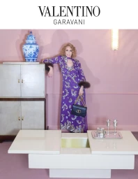 Elderly woman in a purple patterned dress leaning against a cabinet holding a black handbag in a pink interior.