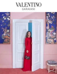 Model in long red gown holding floral handbag standing in a pink room opening a white double door