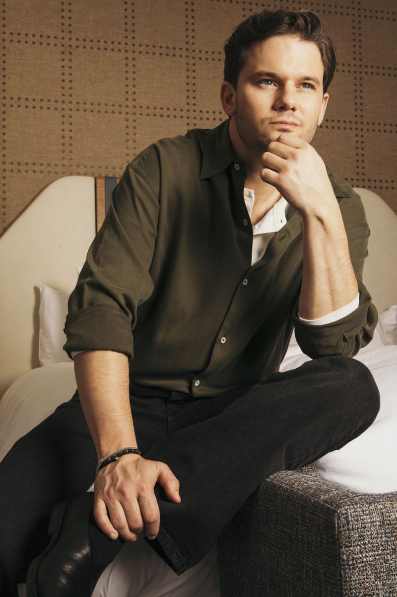 Portrait of a handsome man wearing a green shirt and black pants, sitting on a bed in a neutral colored room with modern décor.
