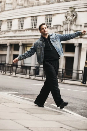 An adult male in a casual outfit walks along a sidewalk in an urban setting with classical architecture in the background on a sunny day.