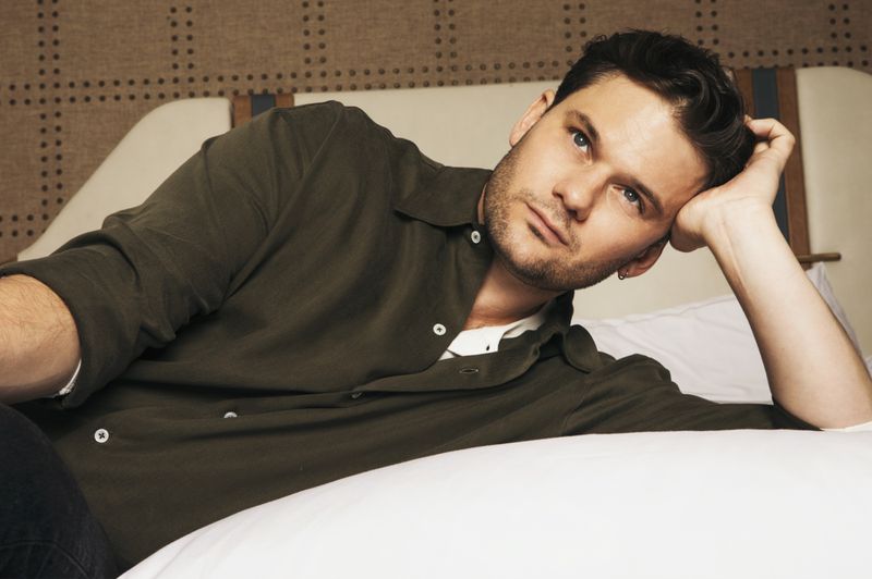 A man in a green shirt lies on a white bed, looking up with a thoughtful expression in a portrait-style, medium shot indoors.