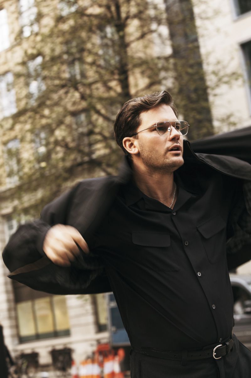 Man in black shirt and coat walks down urban street with blurred motion. Natural light highlights hair. Background building facade.