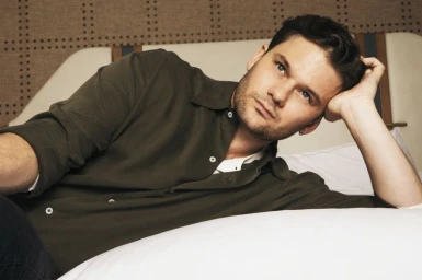 Actor Jeremy Irvine reclining on a white bed wearing an olive green button-down shirt for Grumpy Magazine editorial portrait