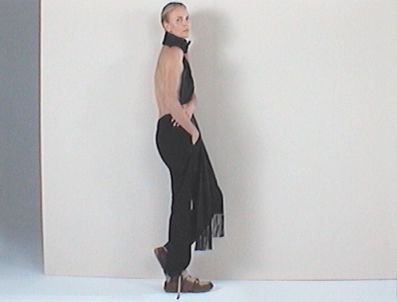Side profile of a fair-skinned adult woman in dark clothing with tan shoes against a neutral backdrop, arms in pockets, one leg raised.