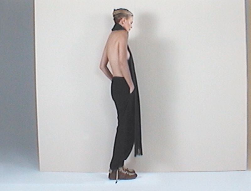 Back view of a woman in dark pants and light brown shoes, wearing a long black scarf, in a photo studio with a neutral background.