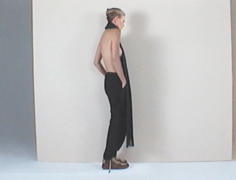 Back view of a woman in dark pants and light brown shoes, wearing a long black scarf, in a photo studio with a neutral background.