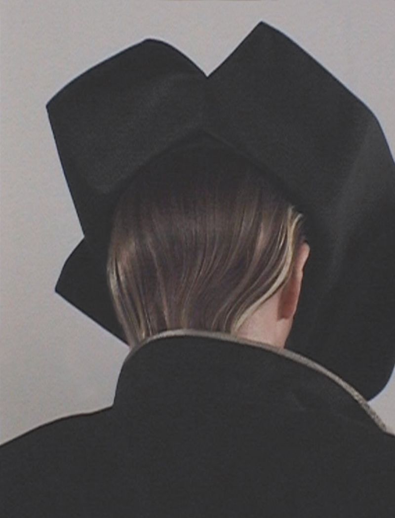 Rear view of a fair-skinned person wearing a black coat and a large black hat against a light gray background in soft lighting.