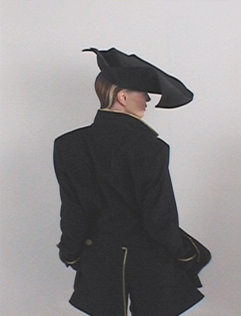 Stylish woman wearing a large, black statement hat and a dark coat with gold trim against a plain background, fashion forward. 