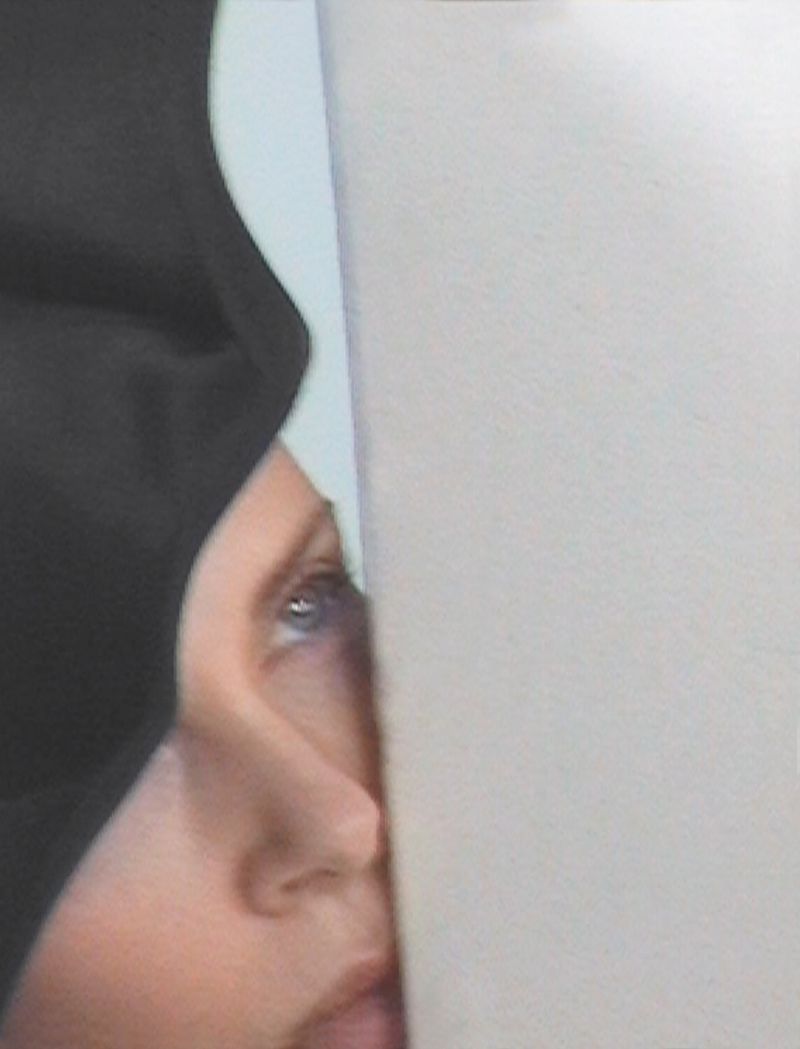 Cropped view of a person with blue eyes partially hidden behind a white wall, wearing black head covering against a light background.