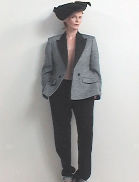 Woman in a gray blazer and black pants stands against a white wall, wearing a black sculptural hat in a studio fashion portrait.