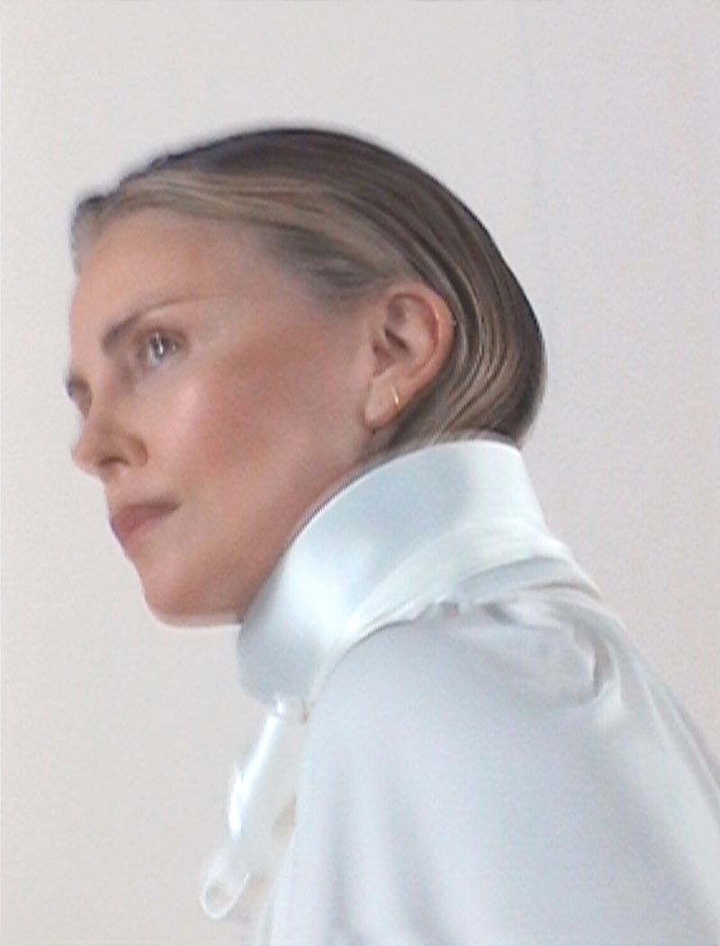 Profile portrait of Charlize Theron wearing a high-collared white garment against a plain backdrop.