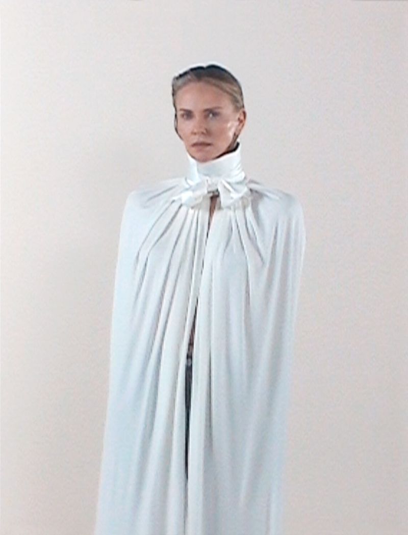 Adult female model in white cloak with high collar and bow, posing in a photo studio against a neutral backdrop, fashion editorial. 