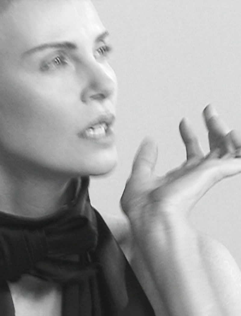Black and white portrait of an adult woman with short hair wearing a dark garment, her hand raised in a gesture against a plain background.
