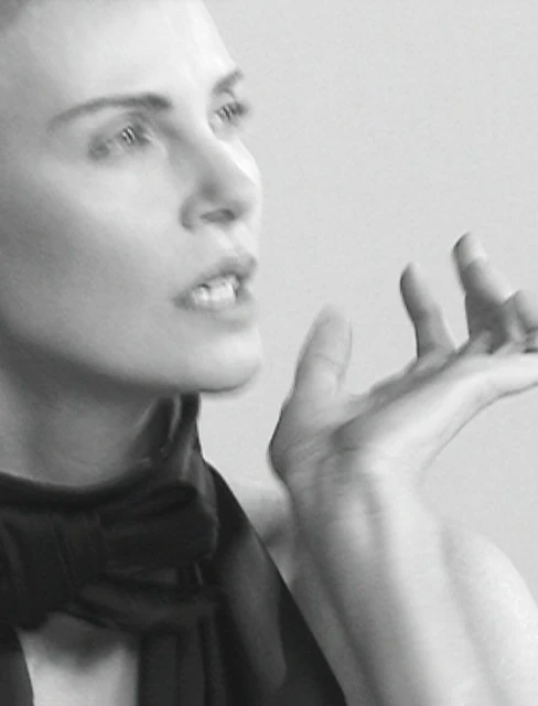 Black and white portrait of an adult woman with short hair wearing a dark garment, her hand raised in a gesture against a plain background.