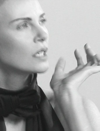 Black and white portrait of an adult woman with short hair wearing a dark garment, her hand raised in a gesture against a plain background.