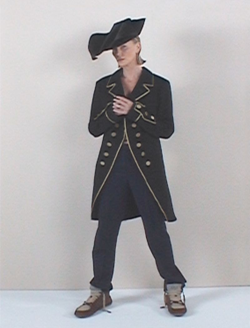 A woman in a black coat and pirate hat stands in a studio, showcasing a unique fashion ensemble with gold trim and brown lace-up shoes.