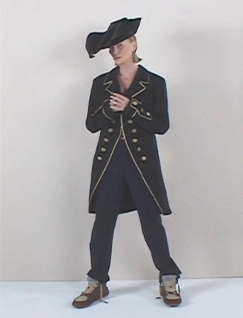 A woman in a black coat and pirate hat stands in a studio, showcasing a unique fashion ensemble with gold trim and brown lace-up shoes.