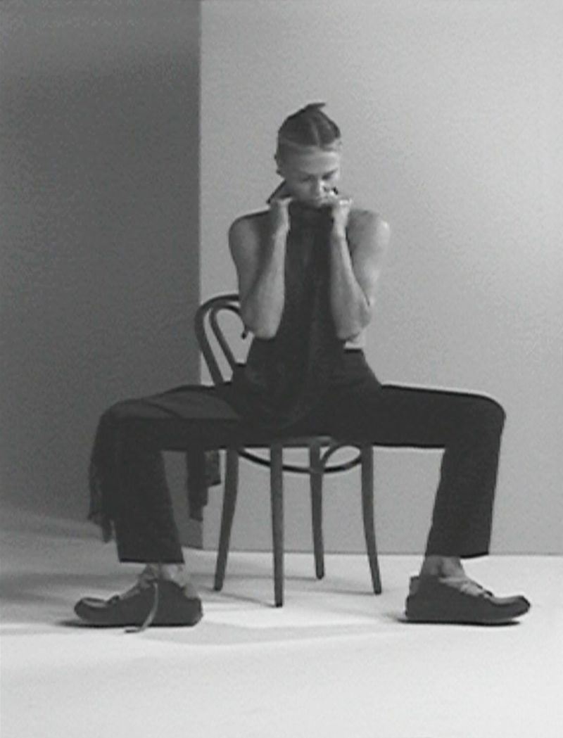Monochromatic fashion shot of a model with braided hair, wearing dark clothing and shoes, seated on a chair against a grey backdrop.