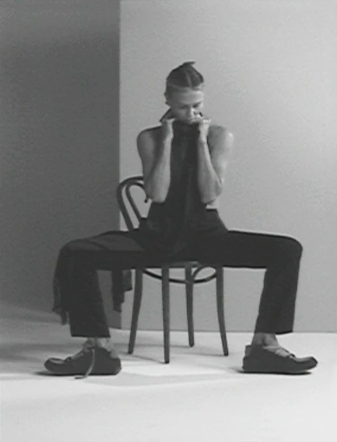 Monochromatic fashion shot of a model with braided hair, wearing dark clothing and shoes, seated on a chair against a grey backdrop.