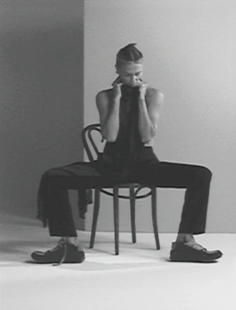 Monochromatic fashion shot of a model with braided hair, wearing dark clothing and shoes, seated on a chair against a grey backdrop.