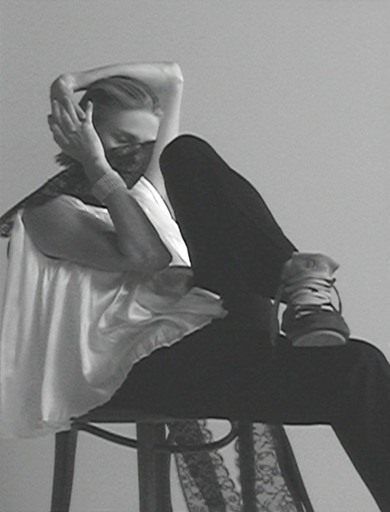 Adult woman in a lace face covering poses while sitting on a chair with one leg extended. White top and dark pants.